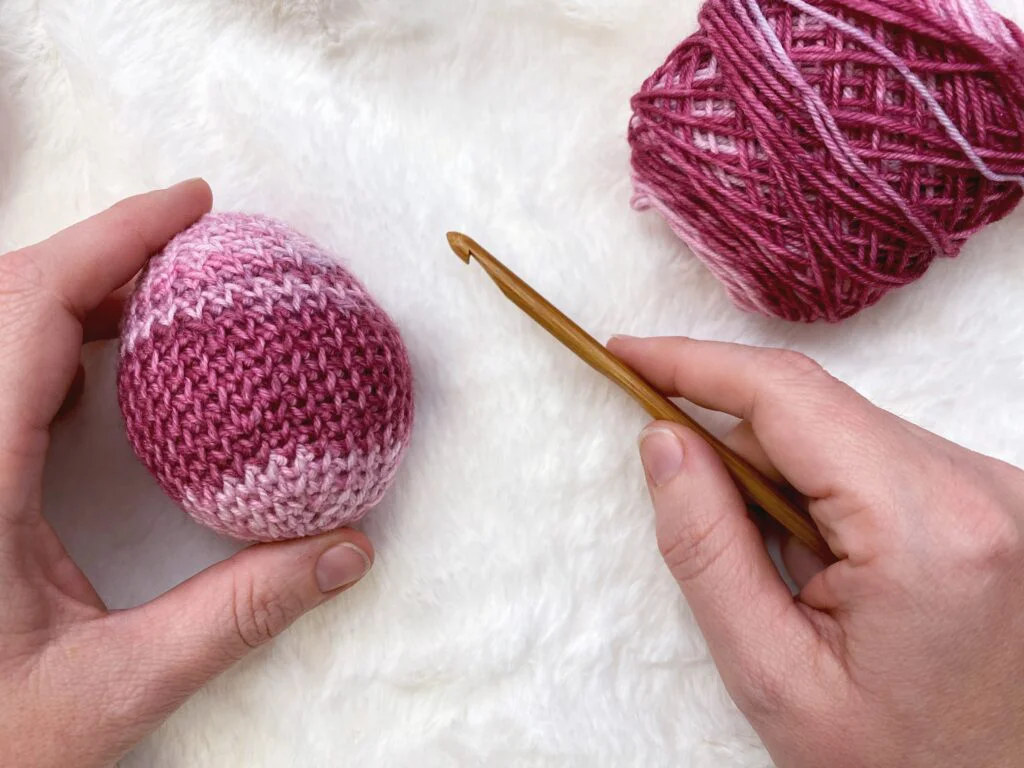 hands holding a crochet hook and crochet easter egg with yarn in background