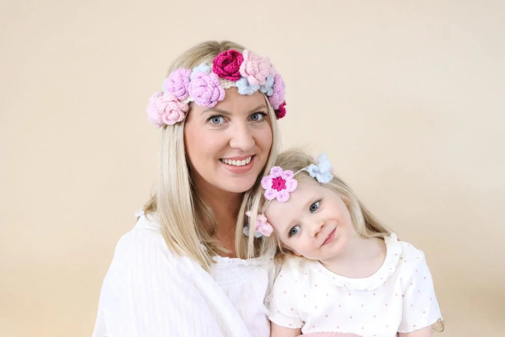 an image of mother and daughter sat infront of a cream backdrop wearing a crochet flower crown each made in pink, purple and pale blue yarn.